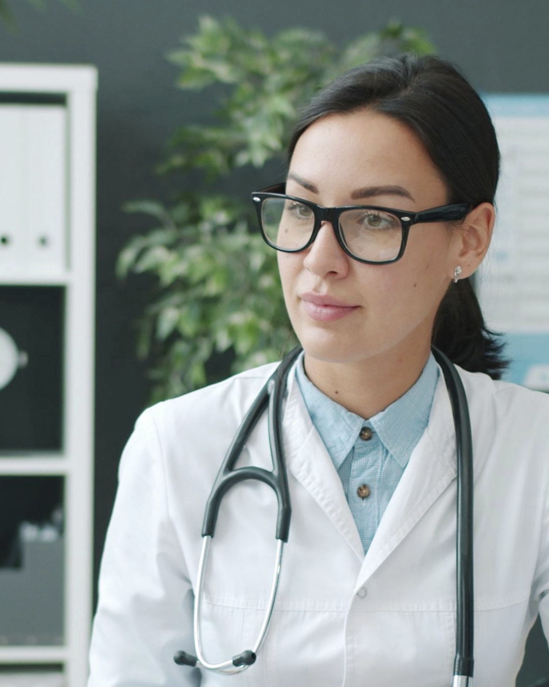 A clinician in a white coat and stethoscope listening attentively during a consultation, in a calm room with plants.